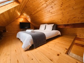 A cozy attic bedroom with a wooden sloped ceiling, a bed with white bedding, a flat-screen TV, and a window.