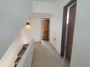 Un couloir se termine par une porte en bois, un luminaire est fixé au mur sur la gauche, et un escalier se trouve sur le côté gauche.
