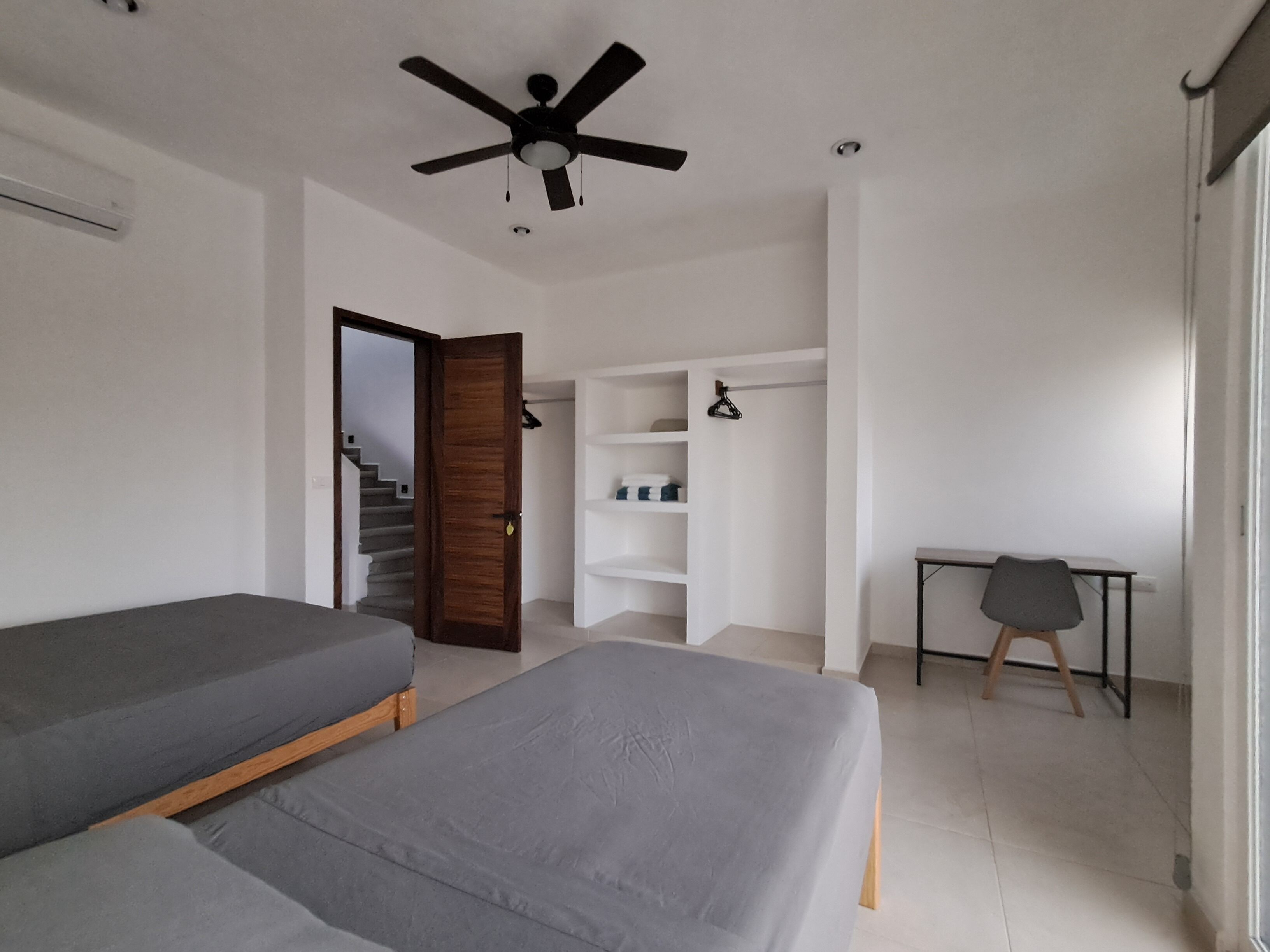 A room with two single beds, a desk, a chair, a wardrobe, a ceiling fan, and a staircase.
