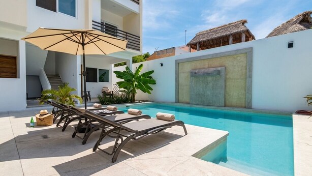 Zona de piscina con sillones reclinables, una sombrilla y una escalera que lleva a un nivel superior.