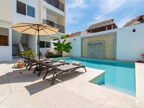Uma área à beira da piscina com espreguiçadeiras, um guarda-sol e uma escadaria que leva a um andar superior.