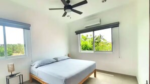 Un dormitorio con cama, ventilador de techo, ventana con vista a follaje y una mesita de noche con lámpara.