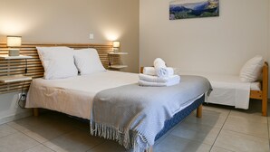Un dormitorio con una cama grande, una cama más pequeña, mesitas de noche, una lámpara de pared y un cuadro de un paisaje.