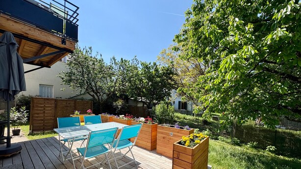 Una terraza de madera con mobiliario exterior, bancales elevados con flores y vistas a una zona residencial.