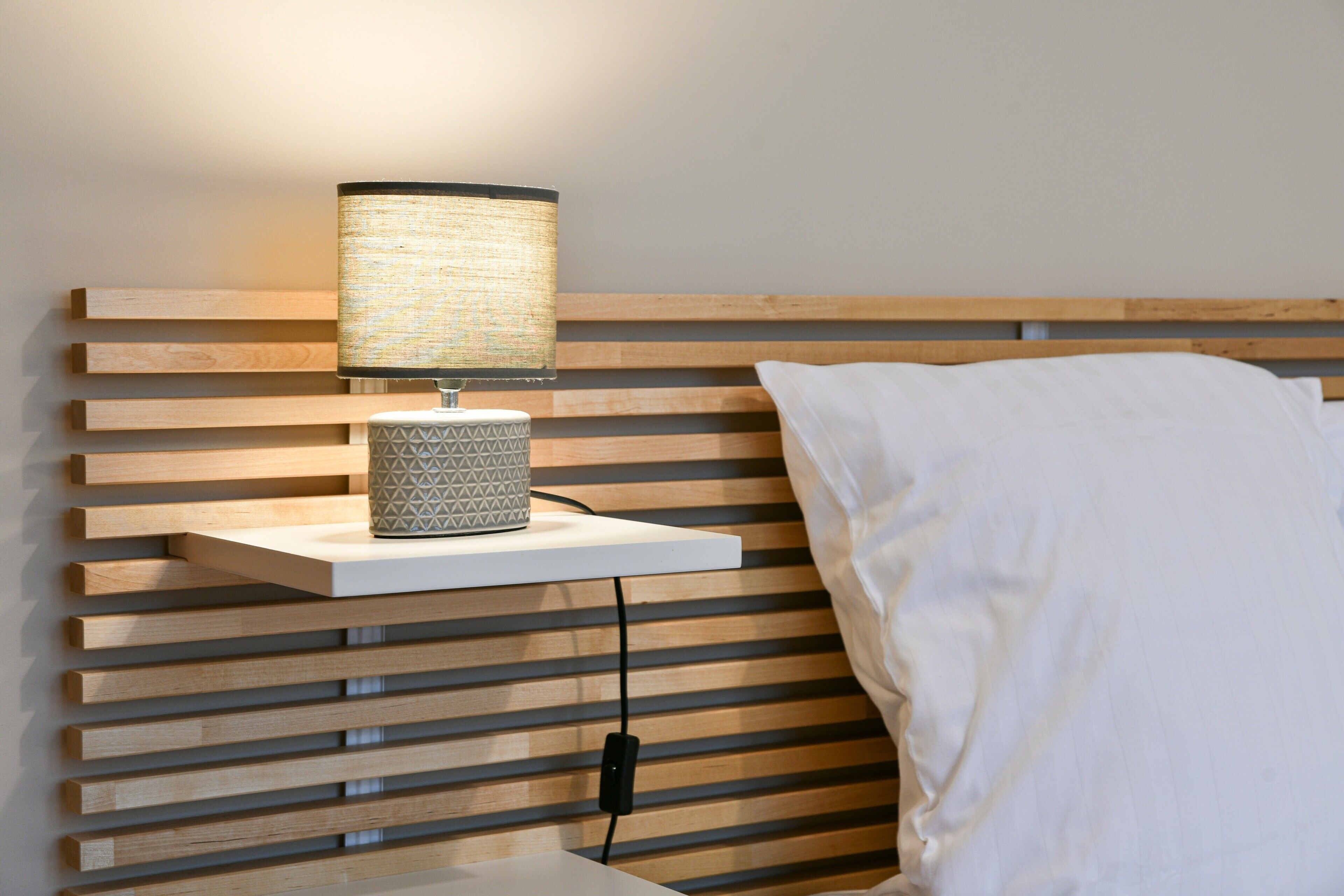 Un lit avec un oreiller blanc, une lampe de chevet posée sur une étagère et une prise électrique au mur.