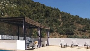 Una piscina con agua cristalina azul, un área de descanso cubierta con sillas y una colina al fondo.