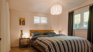 A bedroom with a wooden bed, a bedside table with a plant, a window with curtains, and a hanging light fixture. - Character 3-bdr Newly Reno'd Modern OceanSky Lodge (Britannia Beach)