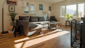 A modern living room with a grey sectional sofa, a glass coffee table, a floor lamp, and a view of the outdoors through large windows. - Character 3-bdr Newly Reno'd Modern OceanSky Lodge (Britannia Beach)
