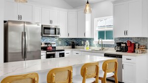 A modern kitchen with white cabinets, stainless steel appliances, a marble island, and wooden chairs. - Brand New Spacious 5BR, 4BA house Built Aug 25, 85 in TV, GameRoom, New Special (Gulfport)