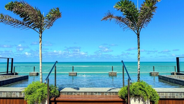 Zona de piscina con vista al océano, escaleras de madera y palmeras.
