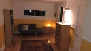 Una habitación pequeña y acogedora con un sofá oscuro, un aparador de madera, una mesita de noche con una lámpara y una estantería.