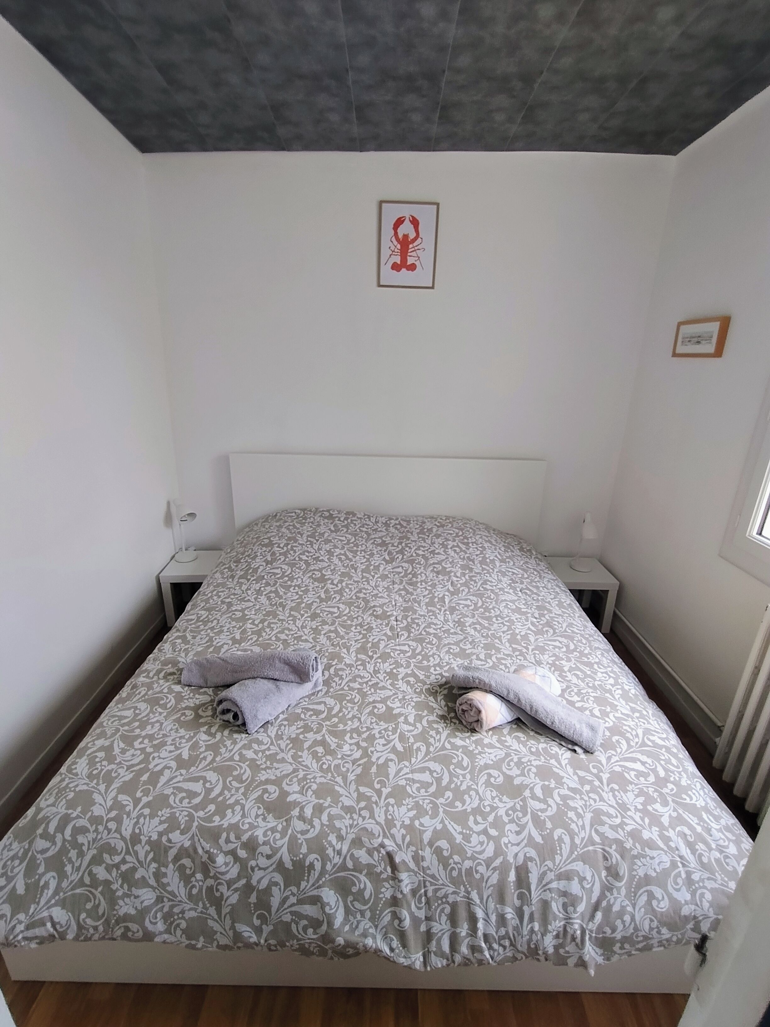 Un llit individual amb un estovat estampat, dues taules de nit, un quadre al mur i una finestra a la dreta.