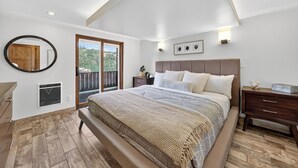 Un dormitorio con una cama grande, suelo de madera, techo con iluminación empotrada, vistas a través de una puerta corredera y una mesita de noche con lámpara.