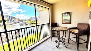 A balcony with a view of a water body and buildings, two wicker chairs, a small round table, and a framed picture on the wall.