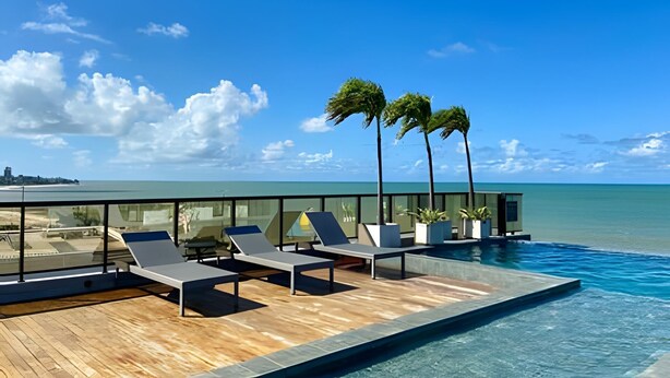 Et rooftop infinity-bassin med liggestole og palmer, med udsigt over en strand og hav.