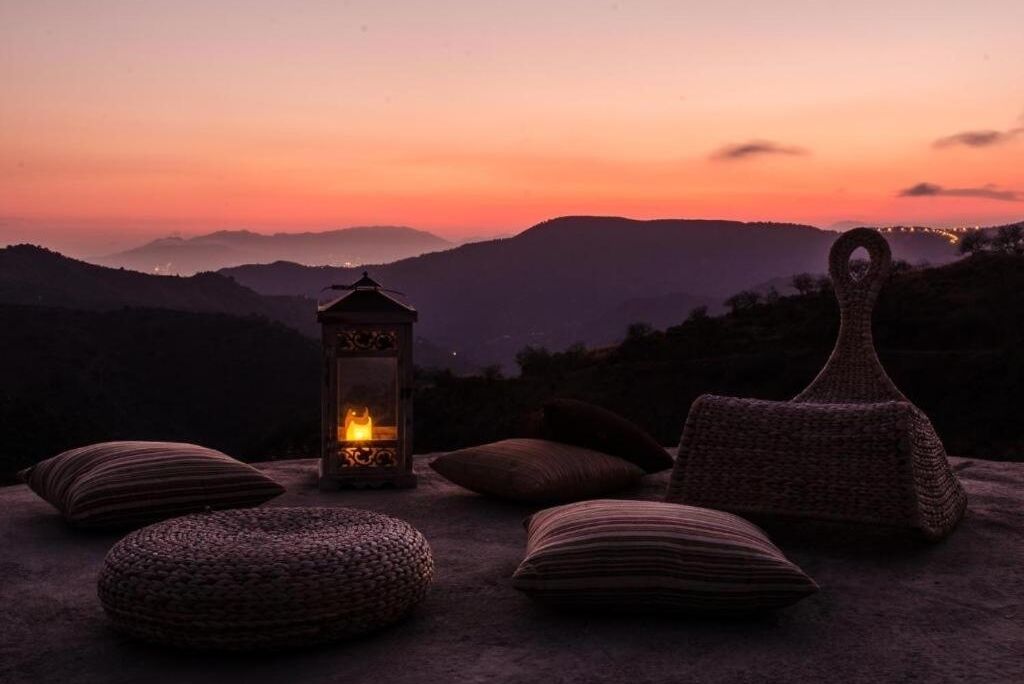 A sunset view with a lantern, cushions, and a woven basket on a stone surface.