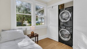 A bedroom with a bed, a nightstand, a lamp, a window with a view of trees and a street, and a wall-mounted washer and dryer. - Downtown Bungalow with Fenced Yard, Walkable to Restaurants, Dog Friendly (Charleston)