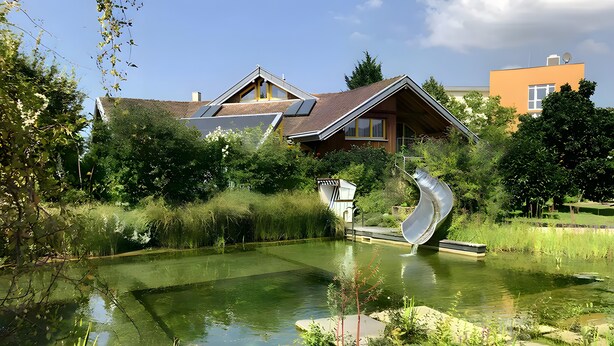 Un étang avec un toboggan, entouré de verdure, et une maison équipée de panneaux solaires.