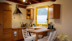 Une cuisine avec des meubles en bois, une fenêtre avec des stores, une machine à café et un coin repas avec des chaises blanches.