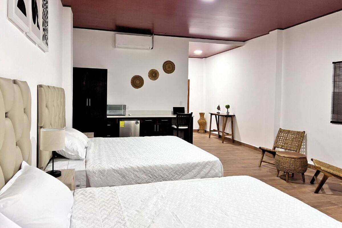 A hotel room with two beds, a kitchenette, a dining area, and a seating area with wicker furniture.