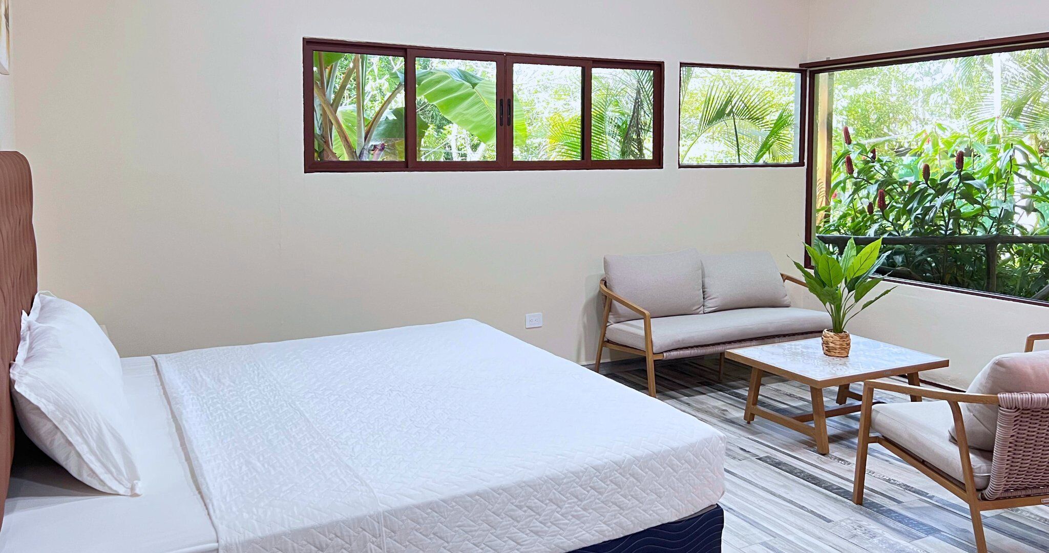 A room with a bed, a sofa, a small table, and a chair. There are three windows with views of greenery.