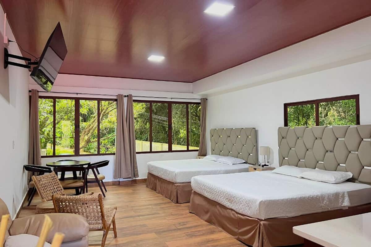 A hotel room with two beds, a dining table, chairs, and large windows offering a view of greenery.