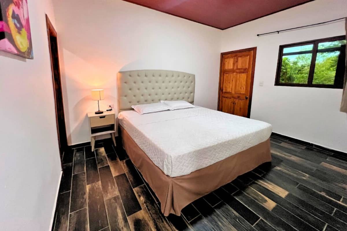 A bedroom with a bed, nightstand, lamp, wooden door, and window.
