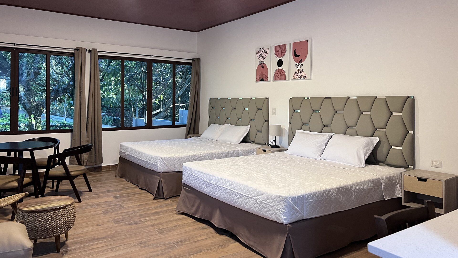 A hotel room with two beds, a dining table, and a view of greenery through large windows.