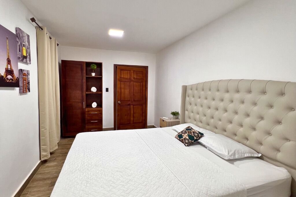 A bedroom with a large bed, a wooden wardrobe, a small bedside table, and a wall with framed pictures.
