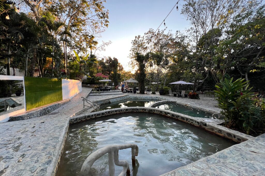 Un espacio exterior tranquilo con una fuente central, senderos de piedra y abundante vegetación.