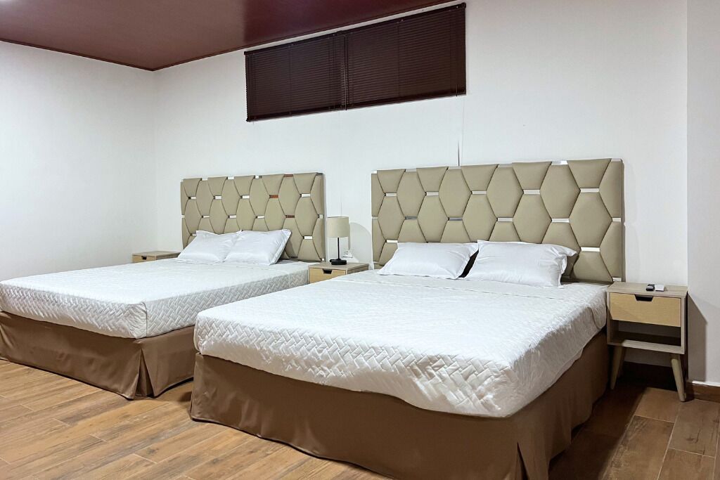 Habitación de hotel con dos camas, cada una con cabecero, mesitas de noche y una ventana con persianas.