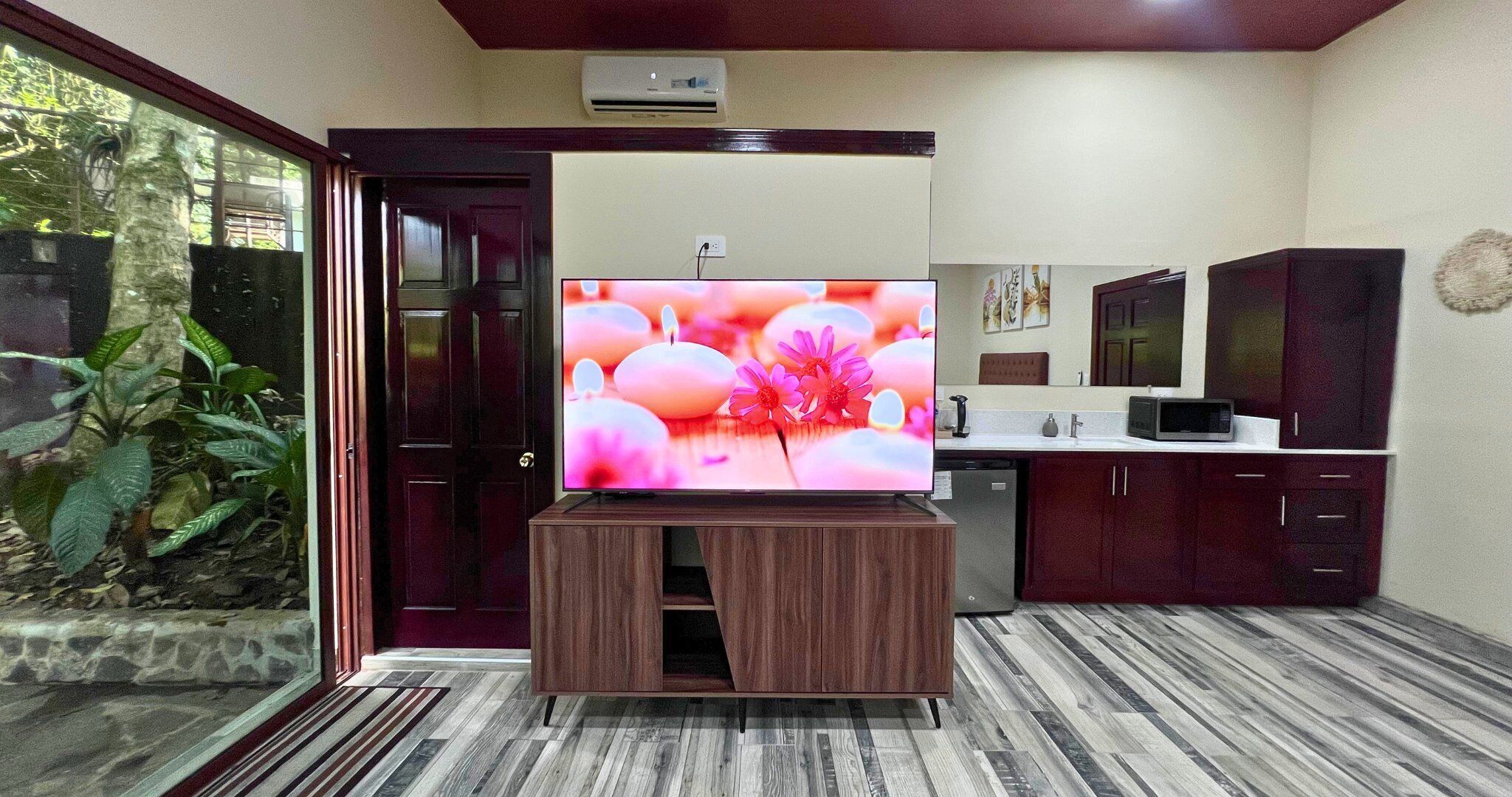 Una habitación con una televisión de pantalla grande en un soporte de madera, una zona de cocina con armarios y un microondas, y un ventanal corredero que da a un jardín con plantas.
