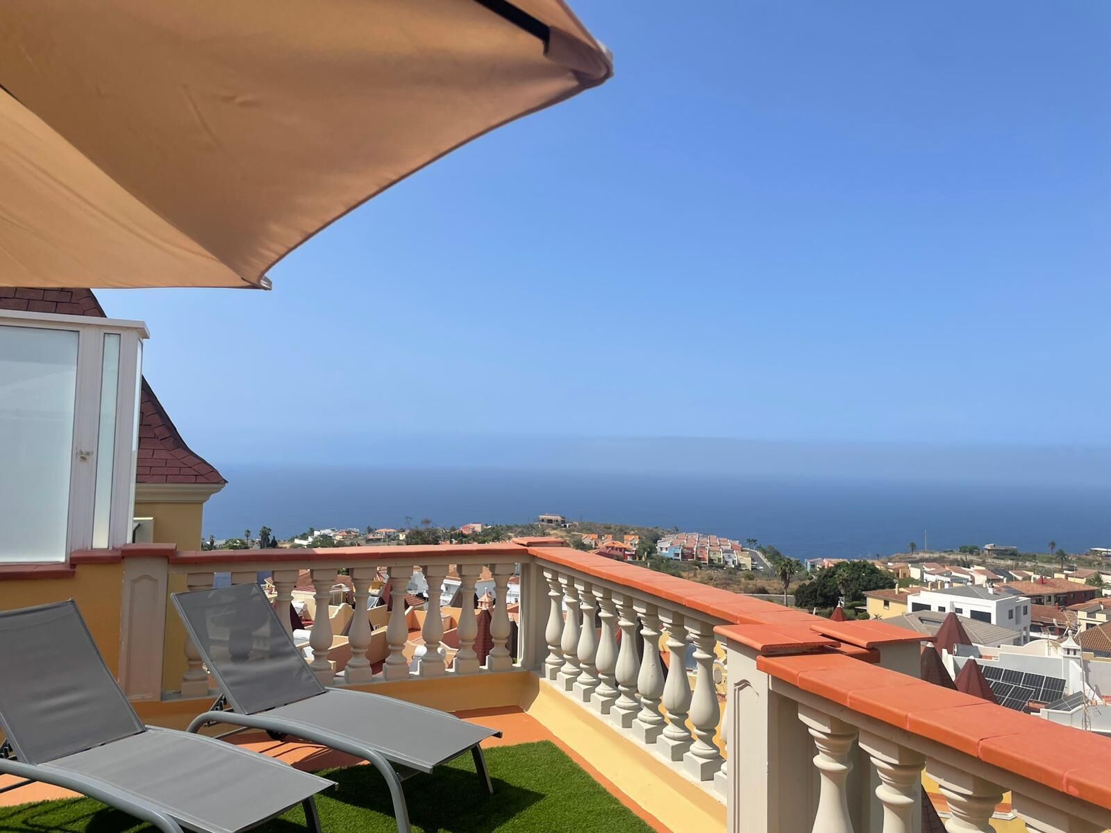 A balcony with two lounge chairs, a beige umbrella, and a view of the sea and town.