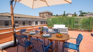 An outdoor patio with a wooden table set for dining, blue chairs, and a large umbrella providing shade. - TreasureRome Forum Rooftop Terrace Colosseum area • 2BR Duplex (Rome)