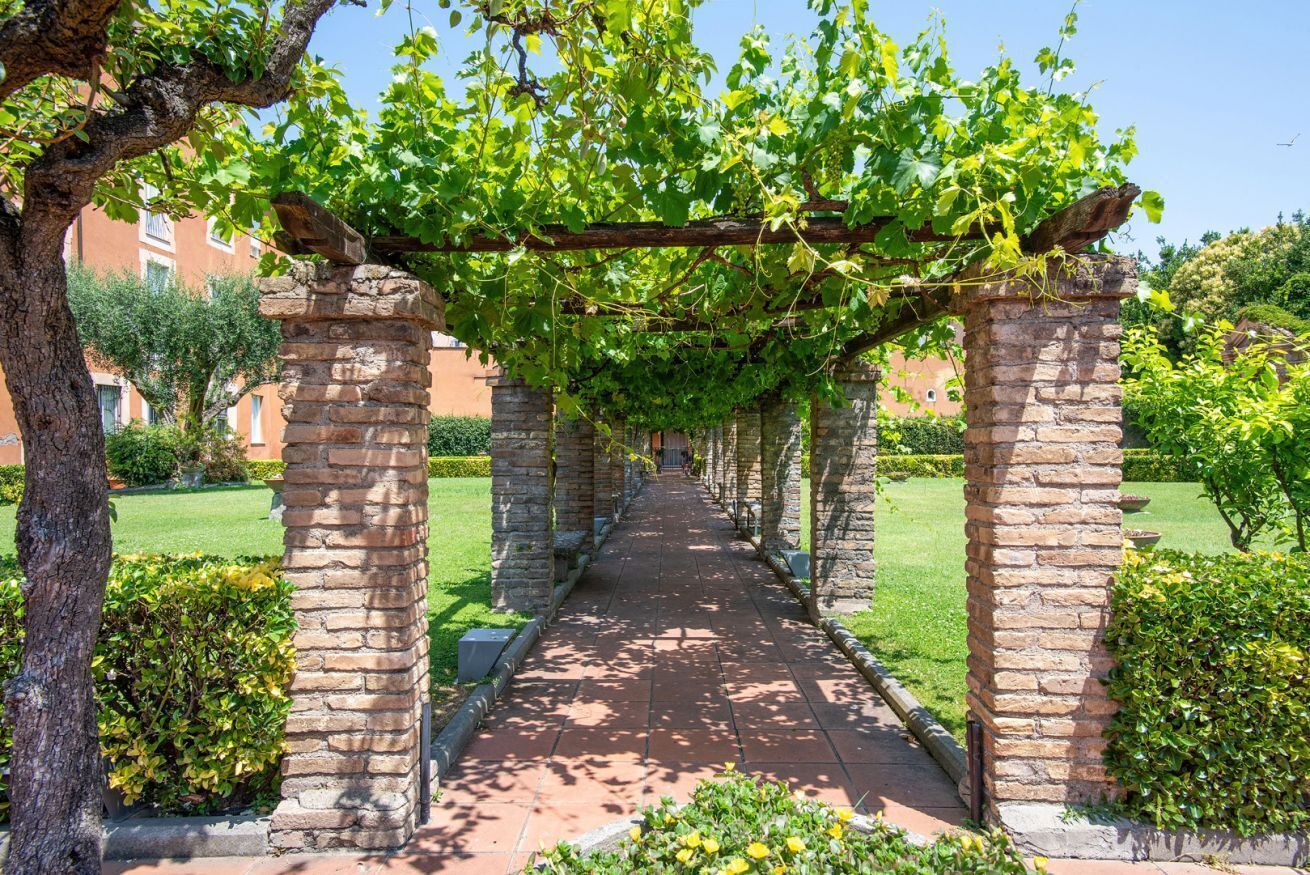 Ein gepflasterter Weg, der von Backsteinpfeilern gesäumt wird, mit einer Pergola darüber und üppiger Bepflanzung.
