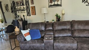 Ein modernes Wohnzimmer mit einem braunen Leder-Modulsofa, einem Couchtisch aus Holz mit Fernbedienungen und einem Essbereich mit weißem Tisch und schwarzen Stühlen.