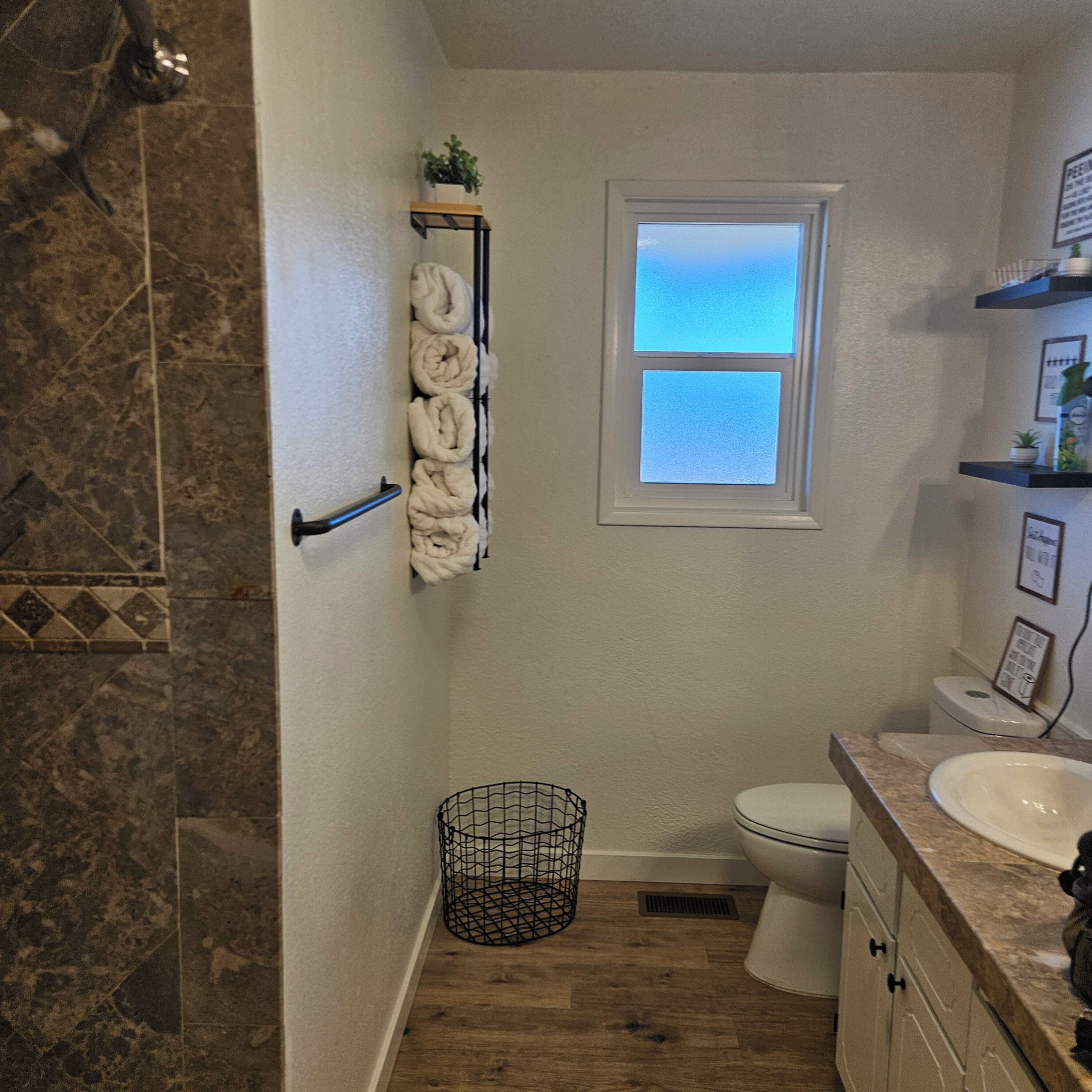 A bathroom with a shower, toilet, and sink. There is a window, a towel rack, and a plant on a shelf.