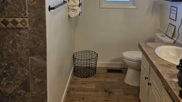 A bathroom with a shower, toilet, and sink. There is a window, a towel rack, and a plant on a shelf.
