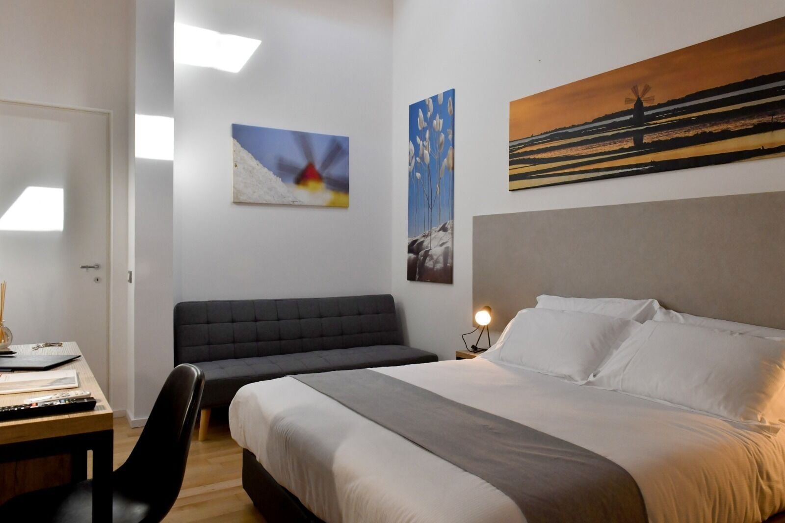 Quarto de hotel com uma cama, uma escrivaninha com cadeira, um sofá e obras de arte nas paredes.