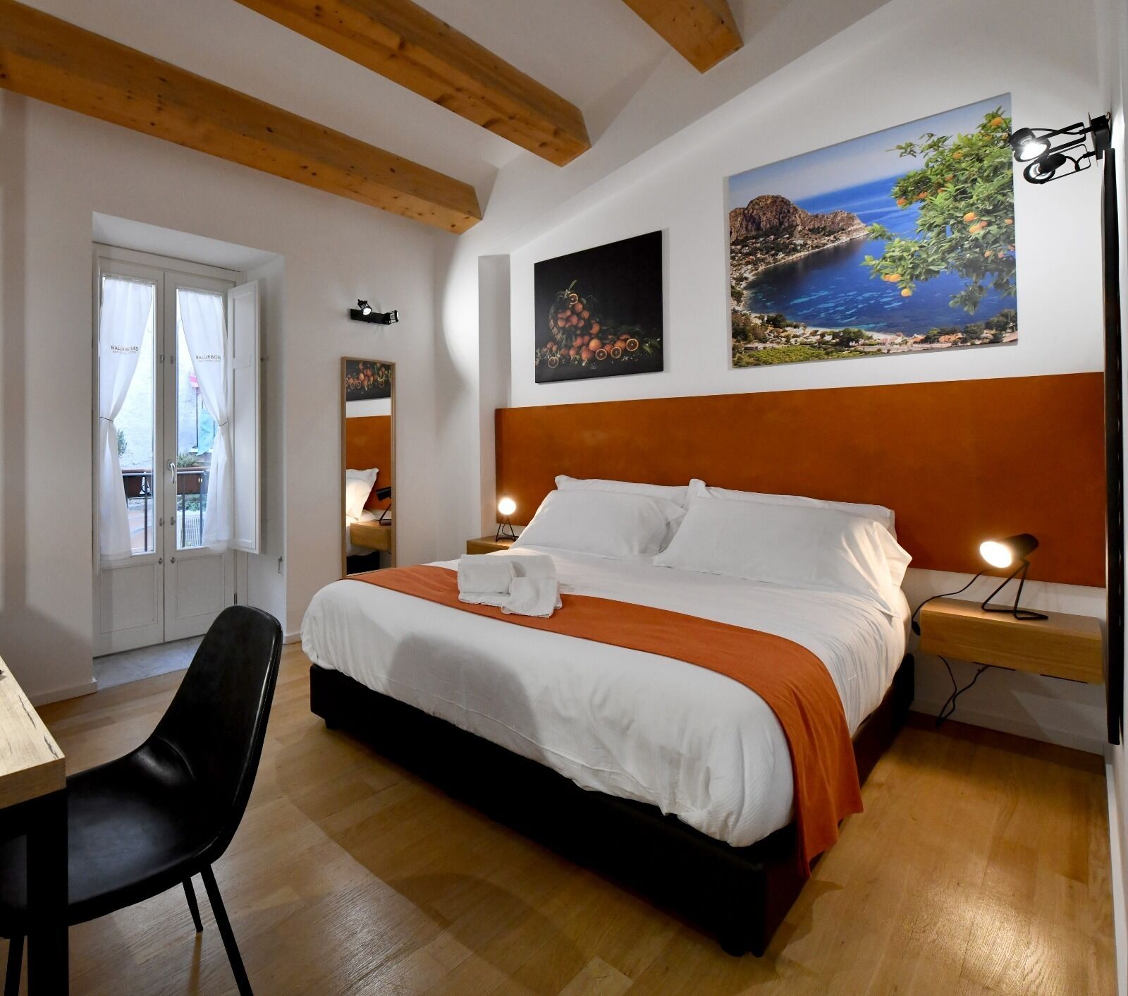 A bedroom with a large bed, a desk, and a chair. There are two paintings on the wall, a window with curtains, and a wooden ceiling.