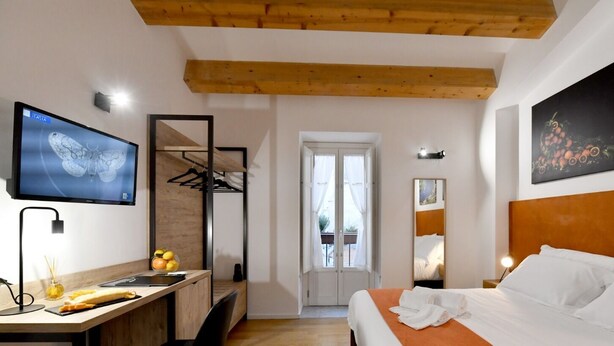 Quarto de hotel moderno com teto de madeira, cama com roupa branca, escrivaninha com computador e um vaso de flores, televisão na parede e uma pintura de frutas.