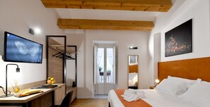 Una camera d'albergo moderna con soffitto in legno, un letto con lenzuola bianche, una scrivania con un computer e un vaso di fiori, una televisione a parete e un quadro con della frutta.