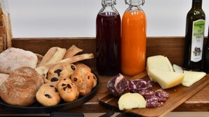 Uma mesa de madeira com diversos alimentos, incluindo pão, doces, queijo e embutidos. Há também duas garrafas de azeite e uma de vinho.
