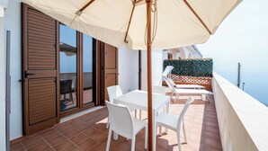 Una terraza con mesa y sillas bajo un toldo grande, una puerta corredera de vidrio y vista al mar.