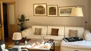 A living room with a sofa, armchairs, a coffee table, and a lamp. There are framed pictures on the wall and a plant.