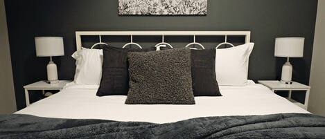 Een net opgemaakt bed met een donkere plaid, witte beddengoed en diverse kussens. Twee nachtkastjes met lampen staan ernaast, en erboven hangt een zwart-wit bloemkunstwerk.