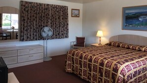 A hotel room with a bed, a desk with a chair, a standing fan, a lamp, and a painting on the wall. - Old Marina Inn (Marina)