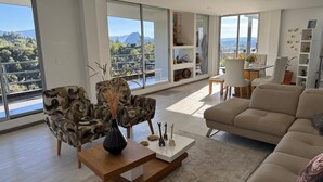 Een ruime woonkamer met een groot raam en uitzicht op een bergachtig landschap. Er staat een moderne bank, een fauteuil met een patroon en een salontafel met decoratieve objecten.