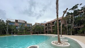 A swimming pool with clear water, surrounded by palm trees and a building with multiple balconies. - Aflora Tulum (Tulum)