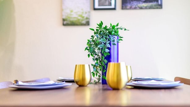 Ein Esstisch mit einer blauen Vase, gefüllt mit grünem Grünzeug, zwei goldfarbenen Gläsern und einem Teller.